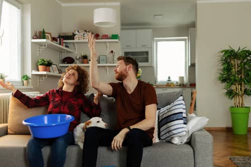 a couple holding a bucket looking at a leak in the ceiling